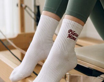 May include: A pair of white ribbed crew socks featuring the text "Pilates Mima" embroidered in dark red thread on the outer side of the ankle. The socks have a classic knit texture and reach mid-calf, providing a clean and simple appearance.