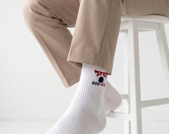 May include: A pair of white crew socks featuring a ribbed texture and a small graphic design on the outer ankle. The graphic consists of a red and white triangular shape above a dark blue circle, with the text ODD WINGS printed in blue and red.