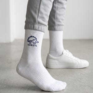May include: White crew socks featuring a dark blue embroidered graphic of a hedgehog above the text CEECIL. The socks have a ribbed texture on the upper portion and are worn with light gray joggers and plain white low-top sneakers.
