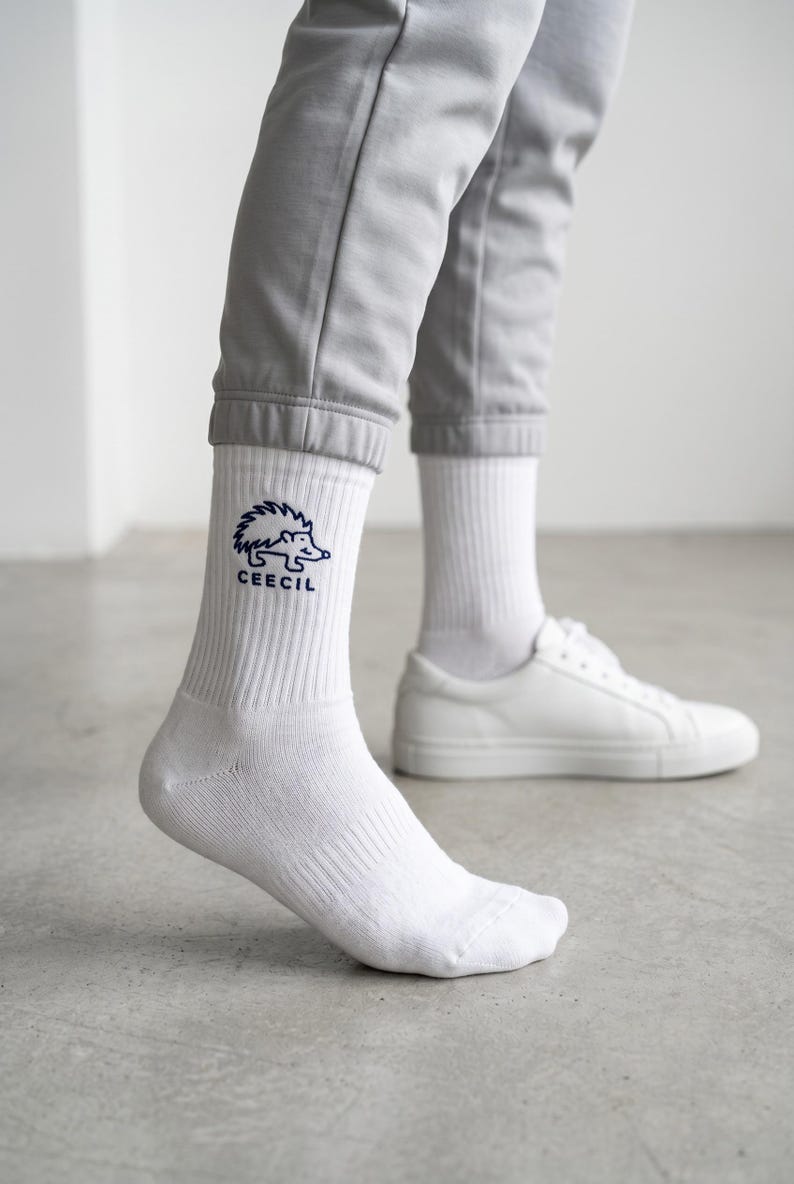 May include: White crew socks featuring a dark blue embroidered graphic of a hedgehog above the text CEECIL. The socks have a ribbed texture on the upper portion and are worn with light gray joggers and plain white low-top sneakers.