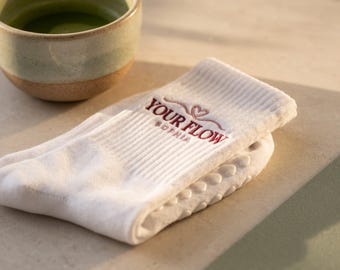 May include: A white crew sock featuring a ribbed cuff with the embroidered text YOUR FLOW and SOPHIA in dark red, accompanied by a small heart icon. The sole of the sock is covered in white silicone grip dots for traction.