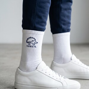 May include: White ribbed crew socks featuring a dark blue embroidered graphic of a hedgehog and the text CEECIL. The socks are worn with white leather sneakers and dark blue pants, providing a clear view of the embroidered design on the ankle.