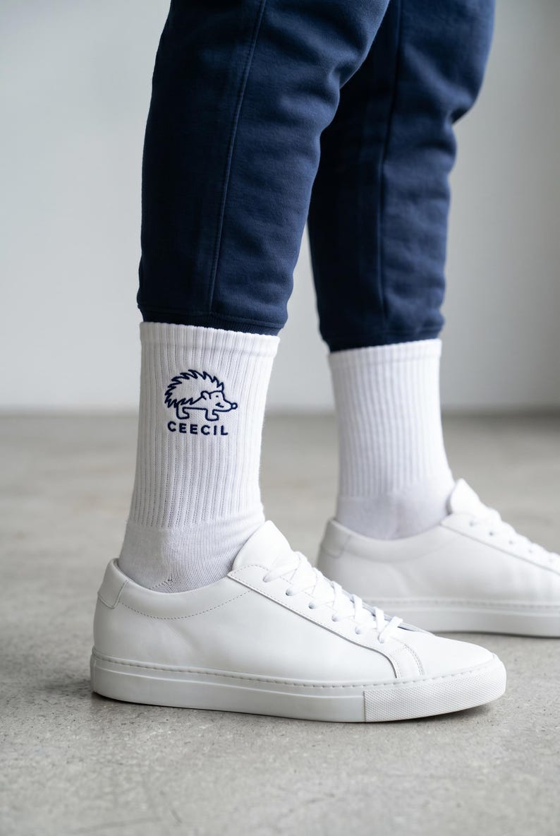 May include: White ribbed crew socks featuring a dark blue embroidered graphic of a hedgehog and the text CEECIL. The socks are worn with white leather sneakers and dark blue pants, providing a clear view of the embroidered design on the ankle.