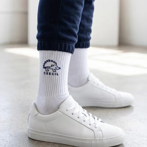 May include: White crew socks featuring a dark blue embroidered hedgehog graphic and the text CEECIL, paired with clean white low-top sneakers and dark navy blue cuffed sweatpants. The socks are ribbed and reach mid-calf in height.