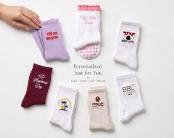May include: A collection of seven crew socks in various colors including white, lavender, maroon, and beige, featuring embroidered text and graphics such as 'Will you marry me', 'My Pilates Studio', 'COFFEE UP!', and 'BBL ALLEGATIONS' with a central text overlay.