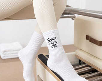 May include: A pair of white crew socks featuring a ribbed texture and black printed text that reads Boozie ATHLETIC CLUB est. 2025. The socks have small white grip dots on the soles for traction, designed for athletic activities or exercise.