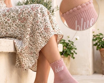 May include: A pair of dusty pink ankle socks featuring a ribbed texture, a scalloped lettuce-edge cuff, and the word "blue" embroidered in white cursive script on the side. The socks are shown in a full view and a circular zoomed-in detail.