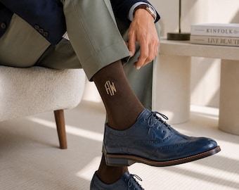 May include: A pair of dark brown socks featuring gold embroidered initials AW, paired with navy blue leather brogue oxford shoes with lace-up closures, decorative perforations, and stacked heels, worn with olive green trousers and a navy blazer.