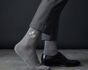 May include: Gray socks with white embroidered initials, paired with gray trousers and black leather dress shoes. The socks feature the embroidered letters "AR". The image is set against a dark gray background.