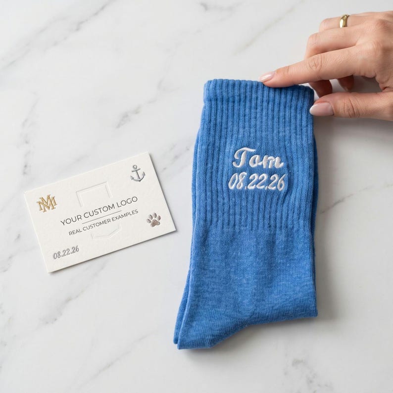 May include: A blue ribbed crew sock featuring white embroidered text that reads Tom and 08.22.26. Beside the sock is a white rectangular card with gold printed text that reads YOUR CUSTOM LOGO, REAL CUSTOMER EXAMPLES, 08.22.26, and icons of an anchor, a paw print, and the letters MM.