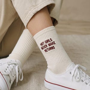 May include: A pair of cream-colored ribbed crew socks featuring the dark red embroidered text "HOT GIRLS NEVER MAKE RETURNS" on the outer side of the ankle. The socks are worn with white canvas sneakers and beige trousers, showcasing a simple design.