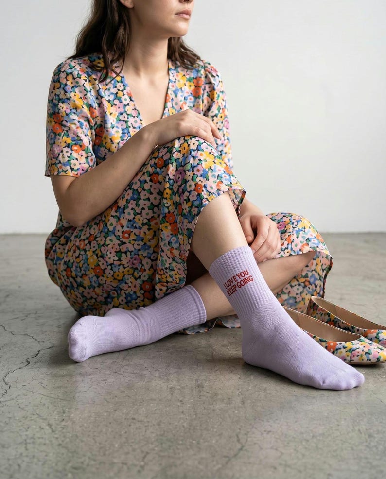 May include: A pair of light purple crew socks featuring the red embroidered text "I LOVE YOU KEEP GOING" on the outer side of the ankle. The socks are made of a ribbed knit fabric and reach mid-calf, providing a simple and clean appearance.