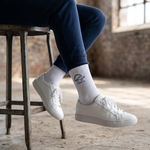 May include: White crew socks featuring a black graphic of a hedgehog and the text CEECIL, paired with white lace-up sneakers and navy blue sweatpants. The socks are ribbed and reach mid-calf, showcasing a clean design with a simple animal illustration.