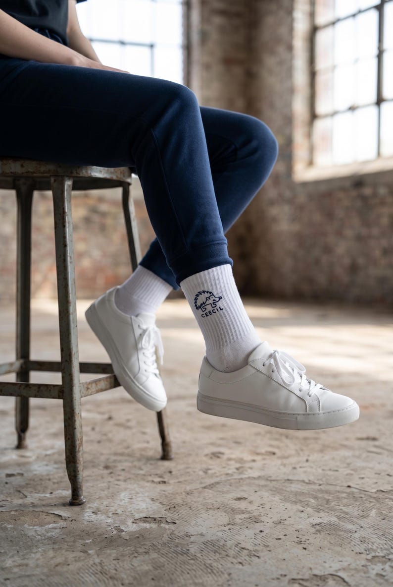May include: White crew socks featuring a black graphic of a hedgehog and the text CEECIL, paired with white lace-up sneakers and navy blue sweatpants. The socks are ribbed and reach mid-calf, showcasing a clean design with a simple animal illustration.