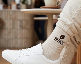 May include: A pair of beige ribbed crew socks featuring a dark brown coffee bean graphic and the text "COFFEE UP!" printed on the side, worn with white leather sneakers and matching beige sweatpants in a casual setting.