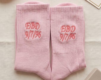May include: A pair of light pink ribbed socks featuring the embroidered text EBD and the date 9/7/96 in a matching pink thread. The socks are laid flat, showcasing the personalized lettering and numbers centered on the upper portion of each sock.