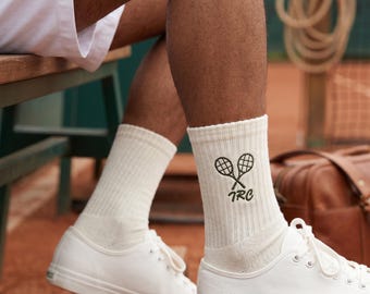 May include: Off-white ribbed crew socks featuring an embroidered design of two crossed tennis rackets in dark green thread above the letters TRC. The socks are worn with white canvas sneakers and white shorts, showcasing a classic athletic style.