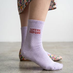 May include: A pair of light purple ribbed crew socks featuring the embroidered red text I LOVE YOU KEEP GOING on the outer side of the ankle. The socks are made of a knit fabric and reach mid-calf, providing a simple and functional design.