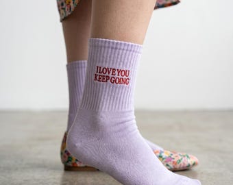 May include: A pair of light purple ribbed crew socks featuring the embroidered red text I LOVE YOU KEEP GOING on the outer side of the ankle. The socks are made of a knit fabric and reach mid-calf, providing a simple and functional design.