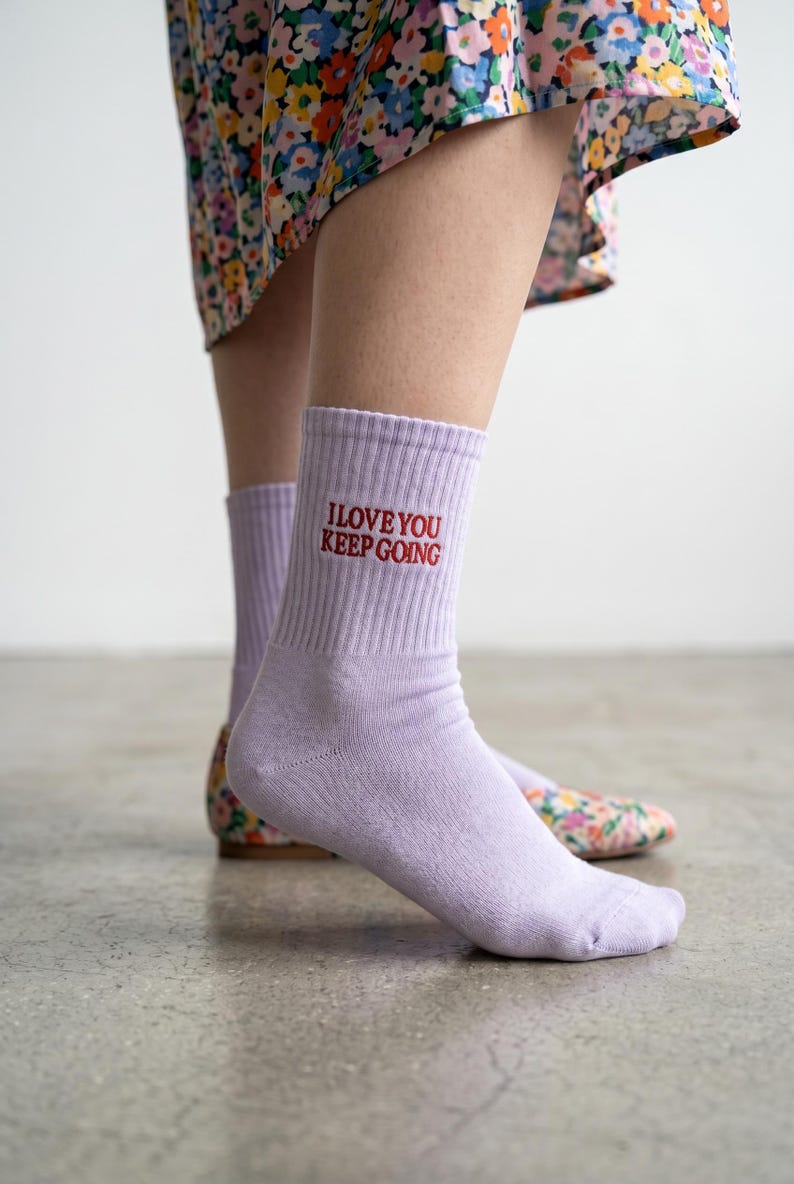 May include: A pair of light purple ribbed crew socks featuring the embroidered red text I LOVE YOU KEEP GOING on the outer side of the ankle. The socks are made of a knit fabric and reach mid-calf, providing a simple and functional design.