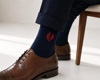 May include: Navy blue socks with red monogram detail, paired with brown leather dress shoes. The socks feature a bold, three-letter monogram. The shoes have a classic lace-up design, and the pants are cuffed.
