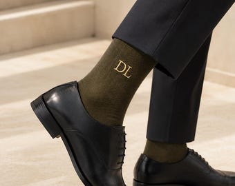 May include: Black leather dress shoes with olive green socks. The socks have the gold initials "DL" embroidered on them. The shoes are laced up and have a polished finish. The person is wearing dark pants.