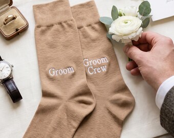 May include: A pair of tan crew socks featuring white embroidered text. One sock displays the word Groom, while the other displays the words Groom Crew. The socks are arranged alongside a beige bow tie, a gold ring in a box, a wristwatch, silver cufflinks, and a white floral boutonniere.
