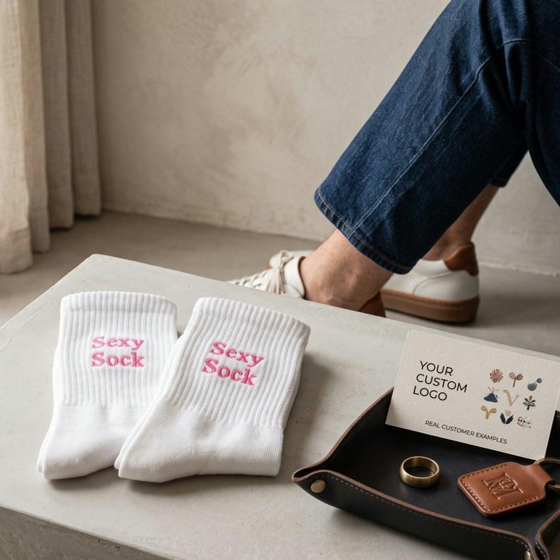 May include: A pair of white crew socks featuring the text Sexy Sock embroidered in pink thread. Next to the socks is a small black tray containing a gold-colored ring, a brown leather keychain, and a card with the text Your Custom Logo.