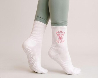 May include: A pair of white crew-length grip socks featuring a pink graphic of two roses tied with a ribbon and the text "MAMAS PILATES" printed on the side. The socks have a ribbed texture and small circular silicone grips on the soles.