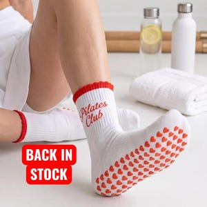 May include: White crew socks with red ruffled cuffs and red heart-shaped silicone grips on the soles. The text "Pilates Club" is printed in red on the side of the ankle. A red rectangular graphic with the white text "BACK IN STOCK" is visible.
