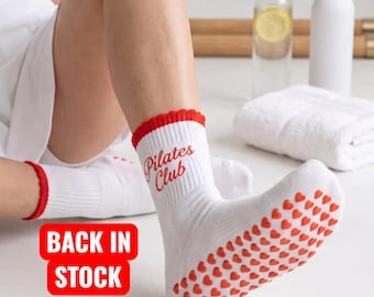 May include: White crew socks with red ruffled cuffs and red heart-shaped silicone grips on the soles. The text "Pilates Club" is printed in red on the side of the ankle. A red rectangular graphic with the white text "BACK IN STOCK" is visible.