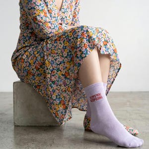 May include: A pair of light purple crew socks featuring the red text I LOVE YOU KEEP GOING printed on the side. The socks are worn with a floral print dress that has a v-neckline and long sleeves, featuring a pattern of small pink, yellow, and blue flowers.