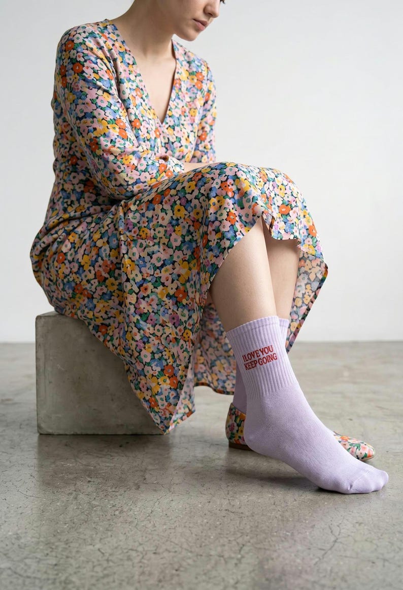 May include: A pair of light purple crew socks featuring the red text I LOVE YOU KEEP GOING printed on the side. The socks are worn with a floral print dress that has a v-neckline and long sleeves, featuring a pattern of small pink, yellow, and blue flowers.
