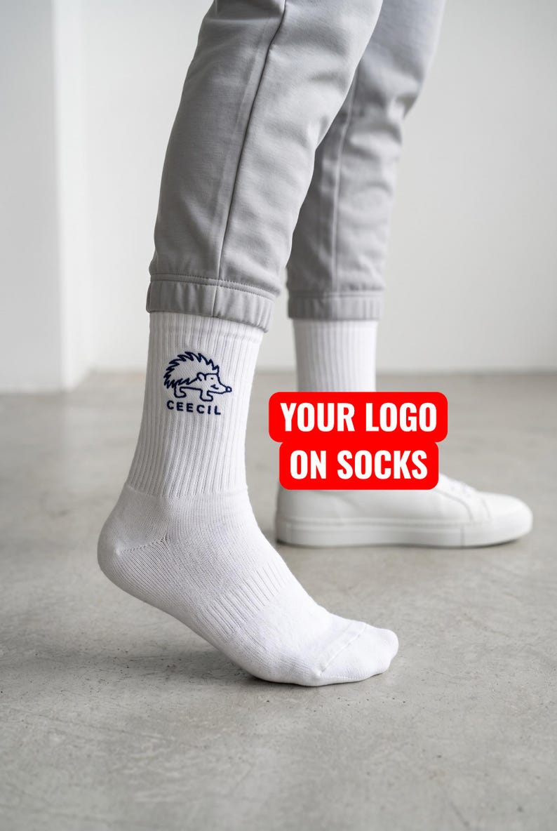 May include: White crew socks featuring a dark blue embroidered hedgehog graphic and the text CEECIL on the outer side. The socks are worn with light gray joggers and white sneakers. A red rectangular graphic with white text reads YOUR LOGO ON SOCKS.