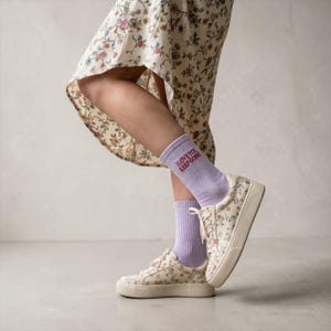 May include: A pair of light purple ribbed crew socks featuring the embroidered red text I LOVE YOU KEEP GOING, worn with floral print low-top sneakers and a matching floral print midi skirt with a high-low hemline and a side slit.