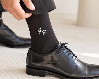 May include: Black dress socks with white embroidered initials. The socks are paired with black leather dress shoes. The image shows a person adjusting the socks, highlighting the personalized detail.