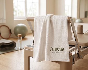 May include: A white cotton towel draped over a beige Pilates reformer machine. The towel features the dark green embroidered text Amelia PILATES CLUB centered on the lower portion. The fabric has a textured terry cloth finish.