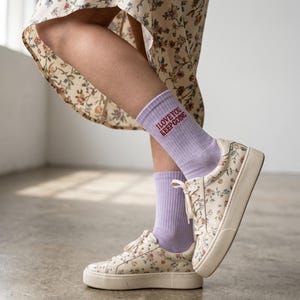 May include: Light purple crew socks featuring the red text I LOVE YOU KEEP GOING, paired with cream-colored low-top sneakers decorated with a small floral pattern in shades of blue, red, and green, and a floral print dress with a high-low hemline.