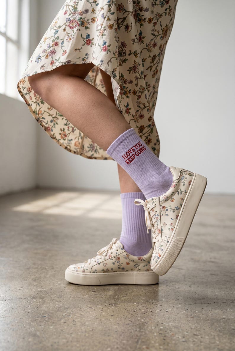May include: Light purple crew socks featuring the red text I LOVE YOU KEEP GOING, paired with cream-colored low-top sneakers decorated with a small floral pattern in shades of blue, red, and green, and a floral print dress with a high-low hemline.