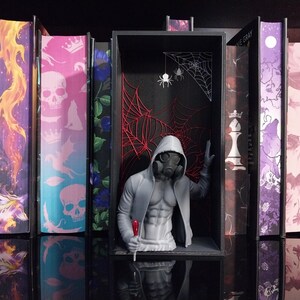 Puede incluir: Un inserto para estanter&iacute;a que presenta una figura gris impresa en 3D con m&aacute;scara de gas y sudadera con capucha, colocada dentro de un marco rectangular negro. El fondo muestra intrincados dise&ntilde;os de telara&ntilde;as rojas y blancas con dos ara&ntilde;as.