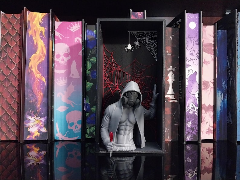 Puede incluir: Un inserto para estanter&iacute;a que presenta una figura gris impresa en 3D con m&aacute;scara de gas y sudadera con capucha, colocada dentro de un marco rectangular negro. El fondo muestra intrincados dise&ntilde;os de telara&ntilde;as rojas y blancas con dos ara&ntilde;as.