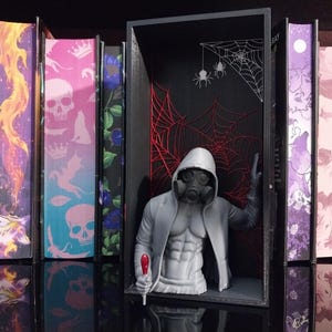 Puede incluir: Un busto gris impreso en 3D de una figura musculosa con sudadera con capucha y m&aacute;scara de gas, colocado dentro de un marco rectangular negro. El fondo del marco presenta ilustraciones de telara&ntilde;as rojas y blancas y dos ara&ntilde;as colgantes.