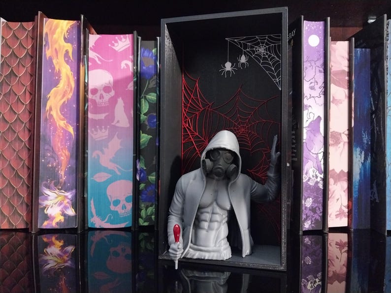 Puede incluir: Un busto gris impreso en 3D de una figura musculosa con sudadera con capucha y m&aacute;scara de gas, colocado dentro de un marco rectangular negro. El fondo del marco presenta ilustraciones de telara&ntilde;as rojas y blancas y dos ara&ntilde;as colgantes.