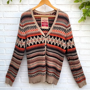 May include: A V-neck cardigan sweater featuring a repeating geometric fair isle pattern in shades of orange, black, and beige. The garment has a button-front closure and a ribbed hem and cuffs. A red and white label is visible at the inner neckline.