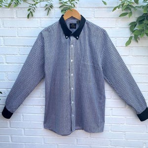 May include: A long-sleeve button-down shirt featuring a blue and white vertical pinstripe pattern. The garment includes a solid black pointed collar and matching black cuffs. A central button placket runs down the front with white buttons.