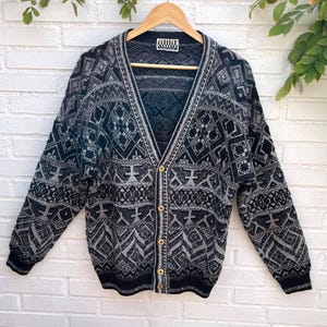 May include: A long-sleeve V-neck cardigan sweater in black, grey, and white with a complex geometric pattern. The garment features a button-front closure with five light-coloured buttons and a ribbed knit hem and cuffs. A label reads College Classics.