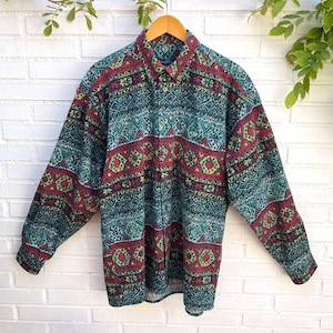 Può includere: Una camicia a maniche lunghe con bottoni caratterizzata da un motivo geometrico ripetuto nei toni del verde acqua, rosso scuro e giallo. La camicia presenta un colletto standard, un'abbottonatura frontale e maniche lunghe con polsini abbottonati.