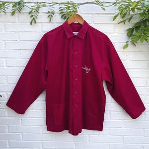 May include: A long-sleeved button-down shirt in a deep burgundy colour. The shirt features a classic collar, a central vertical button placket, and a small embroidered graphic of a bird in flight on the left chest area above the pocket.