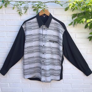 May include: A long-sleeve button-down shirt featuring a light gray front panel with a dark gray horizontal geometric pattern. The collar, sleeves, and back are solid black. The shirt includes a single chest pocket with a black zipper closure.