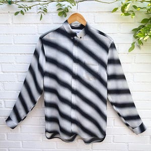 May include: A long-sleeve button-down shirt featuring a diagonal black and white gradient stripe pattern. The shirt includes a standard collar, a single chest pocket on the left side, and a buttoned front placket with a curved hemline.